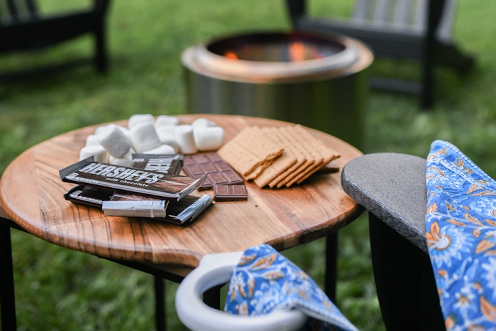 3662CCA2-AFE9-4E1D-A785-1A362A1BC2E8_1_105_c S'mores ingredients on a table with a firepit in the background.